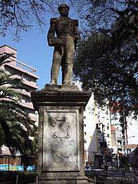 Photograph depicting an urban square, in the center of which is a stone plinth on which is a bronze statue depicting a bearded man in military dress uniform whose right hand holds a bicorn hat