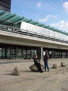 Two people look up at a train stopped at an elevated glass and concrete station