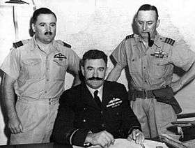Half-length portrait of three military men behind a desk, all with pilot's wings on left breast pocket. One of the men, seated, has a large dark moustache and is wearing a dark winter uniform. The other two, standing on either side of the seated figure, wear short-sleeved tropical uniforms; one of them has a small moustache, the other has a holster on his belt and is smoking a pipe