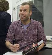 Wolfgang Tillmans, a man wearing a checkered shirt. He is looking up, and is holding a pen over a book as if he was going to sign it.