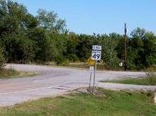 A sign reading "END" above the SH-49 shield. The sign stands next to a road which sharply changes from a well-kept, striped highway to a lower-grade unstriped road at the same place as the sign.