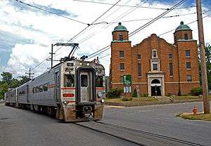 Silver single-level electric passenger train on the street in front of a brick church in Michigan City, Indiana