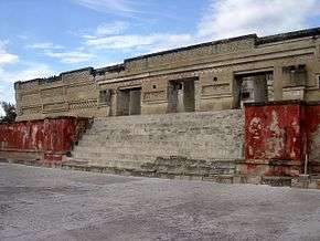 Ruins of a building decorated with geometric patterns in relief. A flight of steps lined by red walls leads to the entrance of the building.