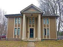 A two-story peaked-roofed wooden house with a projecting front full height porch topped by a pointed roof. It is sided with alternately thick and thin strips of wood running vertically.