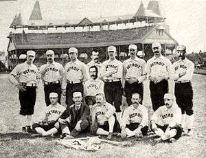 Two rows of men: one row standing behind a second row seated on the ground. The men are wearing white baseball uniforms with "Detroit" across the chest and white baseball caps.