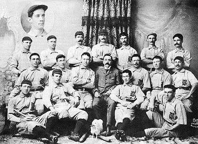 Three rows of men in white baseball uniforms and dark caps; the rear row is standing, the middle row is seated (with a man in a tweed suit in the middle), and the front row is seated on the floor. The baseball uniforms have a dark Old English-style "B" over the left breast.