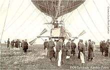 At the top of the picture the lower portion of an airship's envelope can just be seen; mooring ropes reach up to the top edge of the picture; the gondola and propellers can been seen in the centre, surrounded by soldiers, one with his hands in his pockets looking at the camera