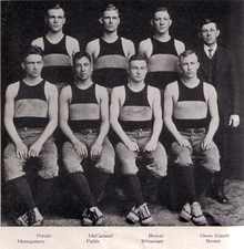 Black-and-white photo showing seven male athletes wearing dark tank tops with a large, light stripe through the middle and a coach wearing a suit and tie.