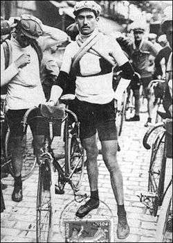 A man standing next to his bicycle; more men and bicycles in the background.