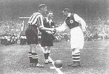 Two football players shake hands in front of a referee over the pitch's center circle.