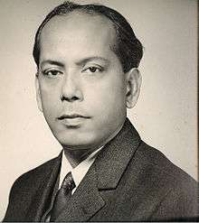 Black-and-white photo of the head and shoulders of a middle-aged man. Brown skin, receding hairline, short dark hair.  He is wearing a suit and looking at the camera with a calm but determined expression.