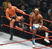 Two men, one Caucasian and one African American, are beside each other and both about to exit a wrestling ring with red ropes. The Caucasian male has light brown hair, and is wearing black wrestling tights, with black wrestling boots and black kneepads. The African American wrestler has dyed blond hair, and is wearing white and blue wrestling tights, and matching wrestling boots with black kneepads. The audience is visible in the background of the photo.
