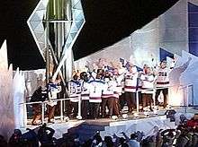 A group of men wearing white hockey jerseys surround a large torch. They light the torch and raise their arms in celebration.