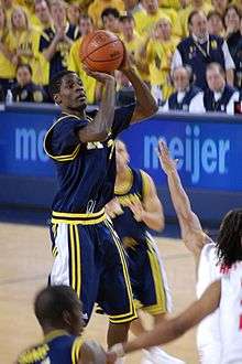A basketball player in a dark blue uniform is shooting a jump shot over the outstretched arm of a defender in white.