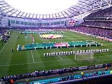 Spectator view of a packed football stadium from behind one of the goals. The opposing stand and part of a lateral stand and the roof structure are seen in the background. A number of people are orderly placed on the pitch, holding banners of several shapes and sizes.