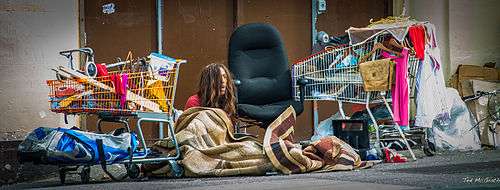 Photograph of a disheveled man sitting amidst shopping carts and junk