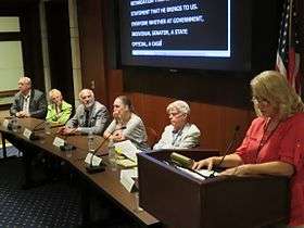 A panel discussion: five people sitting at a long table facing an audience out of frame and the moderator, Janet Albert-Herman, standing at a podium. The room is slightly dark and woody. A screen with captioning is behind the panel.