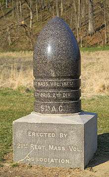 A polished granite monument in the shape of a bullet. The base reads: "Erected by the 21st Regiment Volunteer Infantry." A field and a forest in the background.
