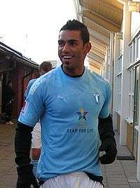 A man wearing a light blue football shirt, white shorts and long black gloves on both hands is seen standing outside a changing room of a football stadium, the man is smiling and looking at something outside the view of the photograph.