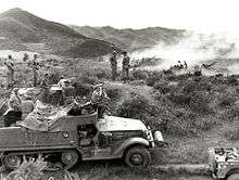 A half track vehicle sits in the foreground in a grassy open area, with a large calibre machine-gun mounted on top. Soldiers using binoculars are sitting on the vehicle, while in the background smoke obscures a number of artillery guns which are firing