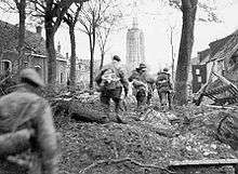 four men in uniform, moving through a tree lined battle damaged street towards a tower in the distance