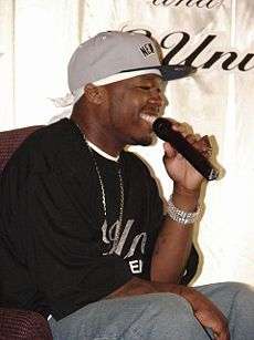 A young black man, seated, wearing a black shirt and a gray baseball cap.  He is smiling and speaking into a microphone.