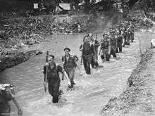 A black-and-white photograph of soldiers marching up a creek. The soldiers have their rifles slung and are knee deep in muddy water