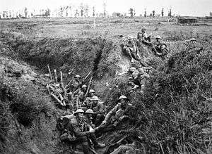 Soldiers wearing helmets and carrying weapons sit in a trench