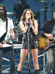 A female teen in a black leather hotpants suit sings into a microphone on a blue-hued stage. Behind her, two electric guitarists and a drummer play their instruments.