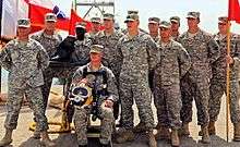Thirteen US Army soldiers are posing for another camera (not seen) with a statuette of their beaver mascot and a red guidon.  One soldier is sitting in front of the rest, wearing a harness and yellow diver's helmet.  To the back-right of the group, a number of flags wave in the wind.