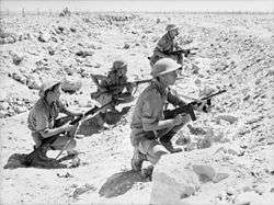 Soldiers crouch behind a low berm amidst a rocky desert area