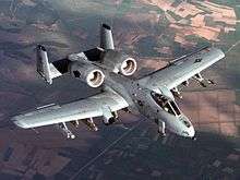 Aerial top view of gray jet aircraft flying above green and brown patchy earth surface. Under each wings are hard points for weapons. The two engines are located aft of the wings and in front of two fin units.