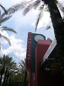 A photograph of a red building with a sign reading "COMMISSARY" in vertical letters all surrounded by palm trees under a blue sky with white clouds