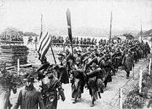 A picture of the American Expeditionary Forces marching across a small bridge holding a flag in France