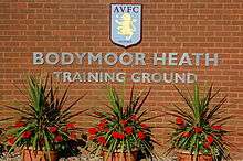 A brick wall with attached lettering reading "Bodymoor Heath Training Ground", surmounted by the AVFC crest