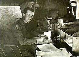 Man in military uniform looking up from a desk covered in papers