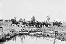 A large formation of soldiers riding horses pass a shallow dam