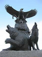 A life-sized bronze statue of an Aboriginal and eagle above him; there is a bear to his right and a wolf to his left, they are all looking upwards towards a blue and white sky