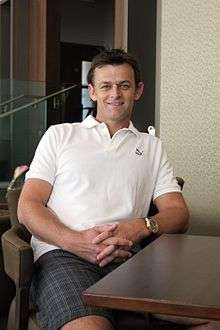 Adam Gilchrist sitting in a chair smiling, leaning on a desk.