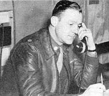 Profile of a white man leaning forward with a large phone handset pressed to his ear. He is wearing a leather jacket over a shirt and tie.
