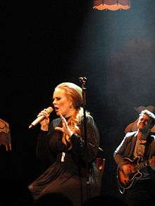 A blonde woman is sitting, while performing and holding her microphone in her hand.