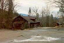 Two unmaintained highways intersect near a large, dilapidated cabin. A second cabin is in the background, reachable by one of the roads. The area is completely surrounded by trees, and large mountains are visible off in the distance.