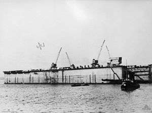 A photo of a rectangular structure with cranes mounted on it floating on a calm body of water. Several boats are visible in front of the structure.