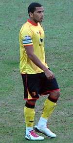 A young man wearing a yellow top and black shorts, standing on a grass field. On his forearm, he is wearing an armband; the word "Captain" is visible.