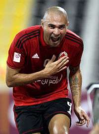 A photograph of a bald Hispanic man in the process of a celebrating a goal, the man is wearing a dark red football shirt and black shorts, he is seen holding his right hand against his chest and with his mouth wide open in the process of cheering.