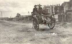 Two men in sombreros riding in a donkey-cart with a line of feet sticking out the back.  They are riding down a dirt street away from the camera, with a line of buildings on the right.  Dated 5/15/1911.