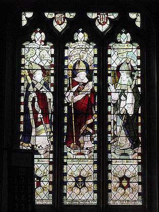 Three stained glass windows, each depciting a mitred and robed figure. All three are carrying staves.
