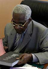 An image of Aimé Césaire in 2003 on a desk reading the cover of a book.