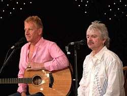 Two man standing at microphones, both are shown facing slightly to the right. Male at left has a guitar and is resting his left arm over it. The second male is shorter and has a moustache.