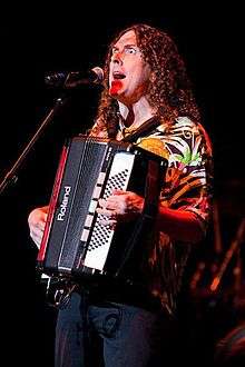 A long-haired man plays an accordion and sings into a microphone.